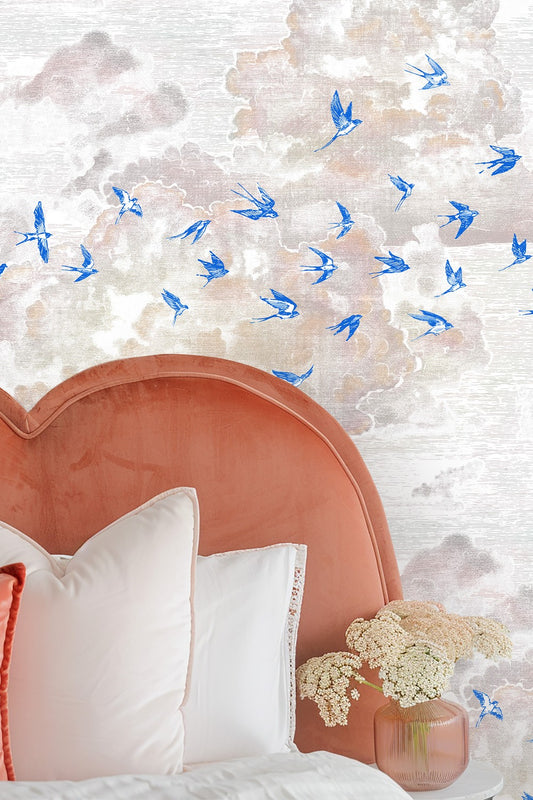 Blue swallows flying across soft pink clouds on a bedroom wall behind a coral velvet headboard